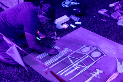 An artist under blacklight painting a glow-in-the-dark artwork on the ground at Art in the Dark