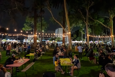 Crowds seated at picnic tables eating under string lights in a tree-lined dining area